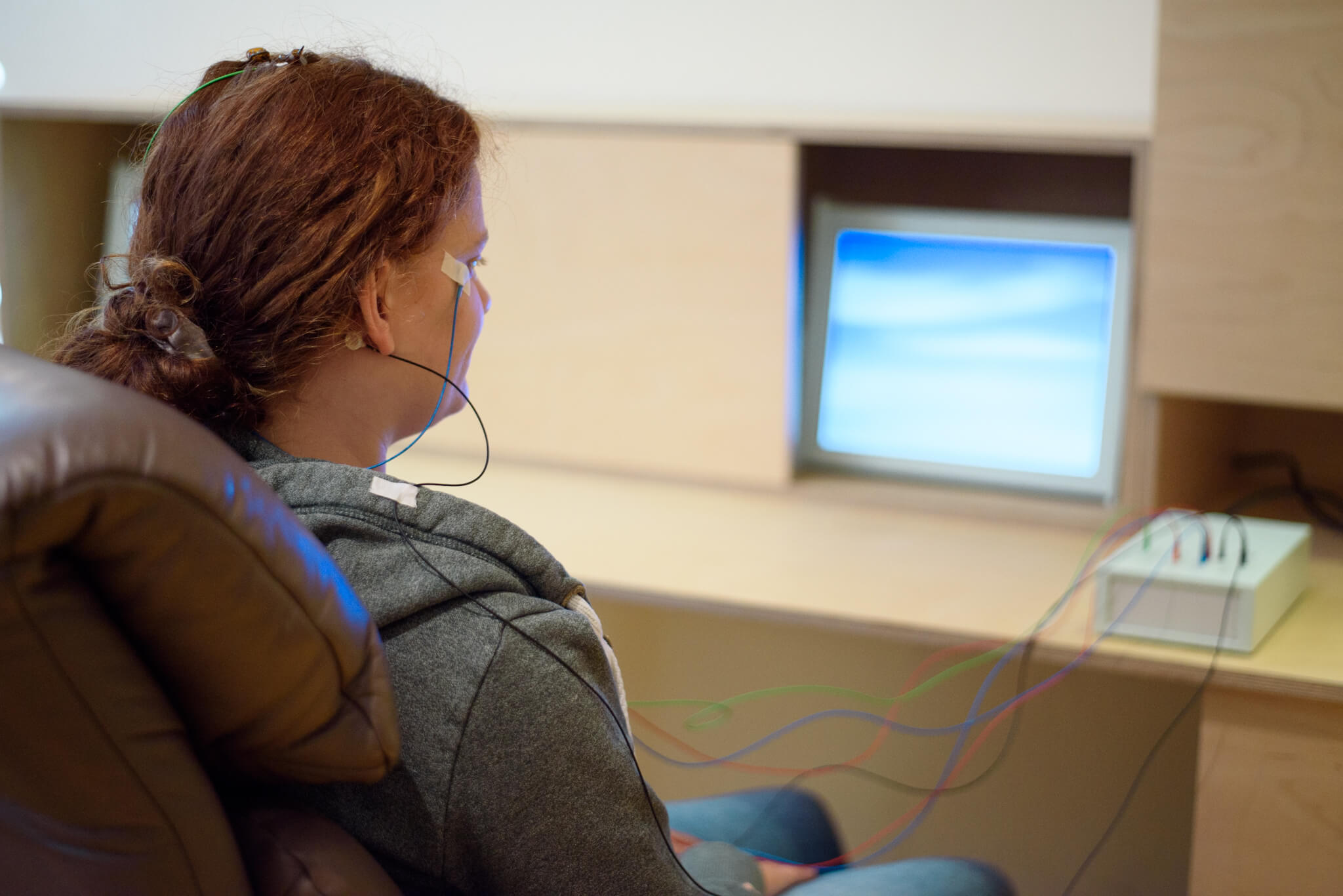 Eine Frau sitzt in einem Sessel vor einem Computerbildschirm. Am ihrem Kopf sind Elektroden befestigt, dere Kabel zu einem hellen Kästchen neben com Computer führen.